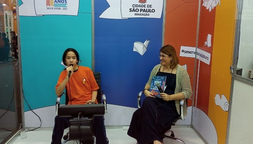 Fotografia mostra uma câmera à frente e atrás duas pessoas sentadas, a esquerda um garoto segurando um microfone e está com a camisa do Imprensa Jovem, ao seu lado esquerdo está Débora Garofalo, ela segura seu livro e está com um macacão jeans e um casaco de tricô, ela segura um microfone. Ao fundo deles é o estúdio do Imprensa Jovem na bienal, podemos ver as paredes adesivadas em verde claro, azul escuro e laranja, também vemos desenhos de livros, uma placa com o logotipo da "Cidade de São Paulo - Educação" e dos "50 anos da sala de leitura", também tem uma placa escrito "#smenabienaldolivro".