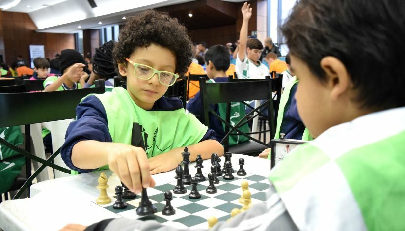 Fotografia em primeiro plano de dois estudantes jogando xadrez, ao fundo mais participantes também jogam com seus pares.