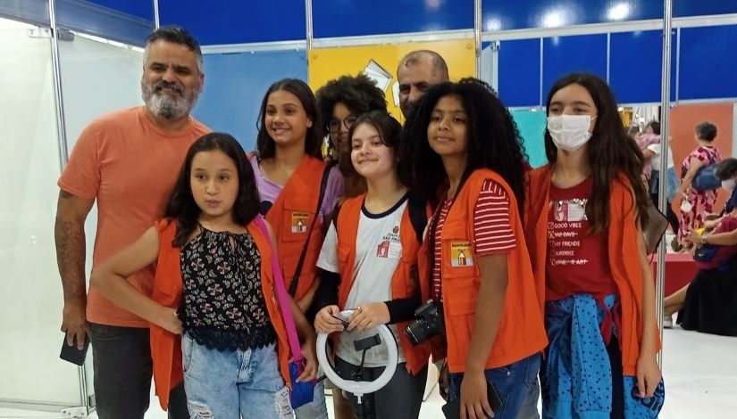 Fotografia mostra 8 pessoas. Da esquerda para a direita, um homem com barba, ele veste uma camisa laranja, calça jeans e segura um celular, ao seu lado tem uma menina com colete laranja do Imprensa Jovem e camiseta rosa; em seguida uma menina com óculos e um homem (ambos só mostra uma parte do rosto), a frente dessa menina tem outra menina com um colete laranja, camiseta do uniforme da rede municipal de ensino, ela segura uma ring light, ao seu lado tem uma menina com o mesmo colete, camiseta vermelha com listras brancas, uma câmera fotográfica no pescoço e segura um celular, por último tem uma menina com o mesmo colete, camiseta vermelha e uma blusa de frio na cintura na cor azul com estrelas pretas. Ao fundo deles vemos paredes na cor azul escuro.