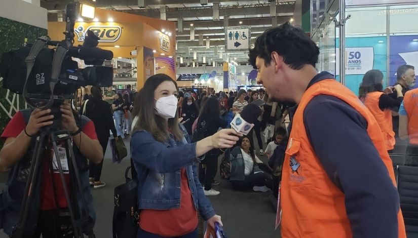 Fotografia mostra à esquerda uma pessoa segurando uma câmera de vídeo que está sobre um tripé. à sua frente está uma mulher segurando um microfone e quem fala ao microfone é um integrante da Imprensa Surda, ele está de perfil, de colete laranja e blusa azul escuro.