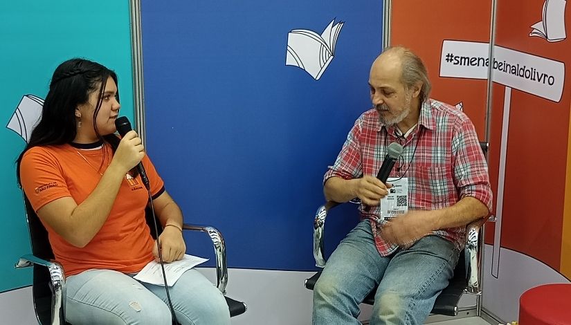 Fotografia mostra duas pessoas sentadas; à direita está Odilon Moraes, ele segura um microfone e está de camisa xadrez e calça jeans; à esquerda está uma menina também segurando microfone, ela está com a camiseta laranja do Imprensa Jovem e calça jeans. Ao fundo deles é o estúdio do Imprensa Jovem na bienal, podemos ver as paredes adesivadas em verde claro e azul escuro, a que está atrás de Odilon é laranja e vemos o desenho de uma placa com o texto "#smenabienaldolivro".