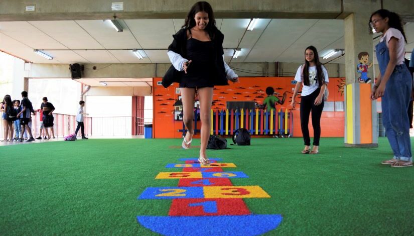 Menina está pulando amarelinha. Ao seu lado esquerdo há duas meninas em pé que estão a observando. À sua direita há um grupo de crianças conversando.