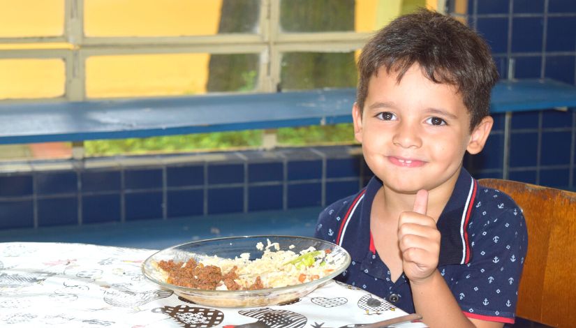 Fotografia de um menino sentado à mesa com seu prato de refeição com arroz, feijão, folhas verdes e carne. A criança veste camisa polo azul marinho com pequenos símbolos de âncora. Ele olha para a câmera sorrindo e fazendo sinal de positivo.
