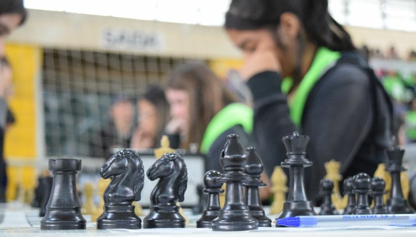 Fotografia de um tabuleiro de xadrez e as peças pretas em primeiro plano, ao fundo desfocado aparecem jogadoras diante dos tabuleiros disputando uma partida.