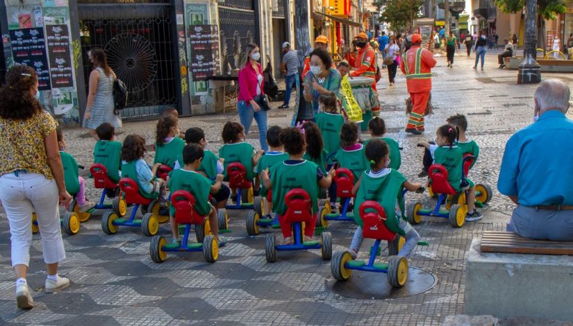 Grupo de crianças andando com suas motocas no centro de São Paulo. As crianças usam coletes verdes. A frente do grupo a professora conversa com elas e movimenta as mãos como se fosse bater palmas. Ao redor do grupo há trabalhadores e pedestres que caminham pelo local.