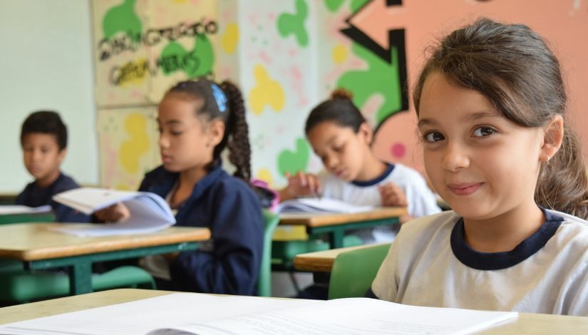 Quatro estudantes estão sentadas em suas carteiras, há um livro na frente deles. Uma menina aparece à frente da fotografia, os outros estão desfocados mais ao fundo.