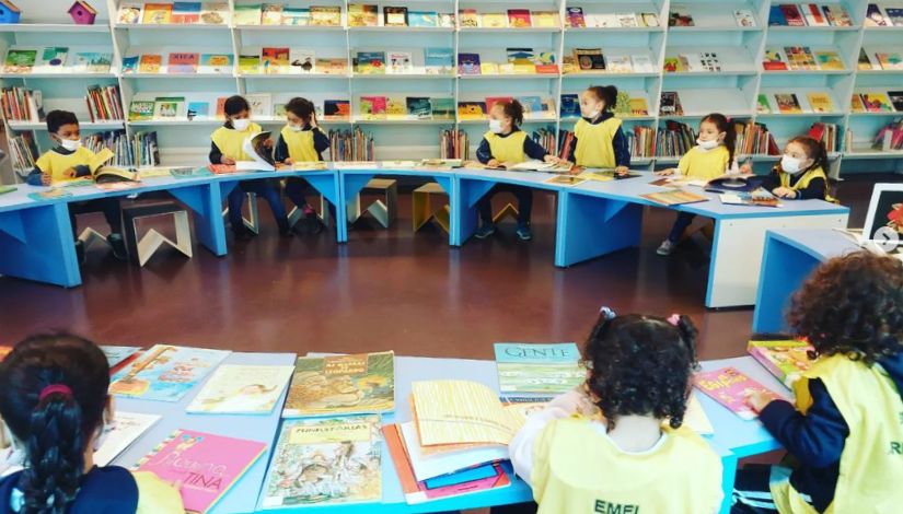 Crianças sentadas em bancos atrás de mesas dispostas em círculo. Sobre as mesas vários livros. Ao fundo há estantes de livros em toda parede.