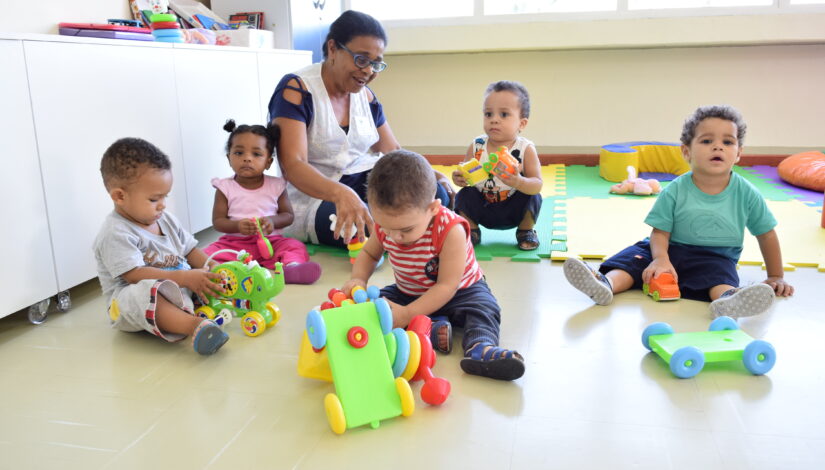 Professora de Educação Infantil sentada no chão da sala com cinco bebês sentados a sua volta brincando.