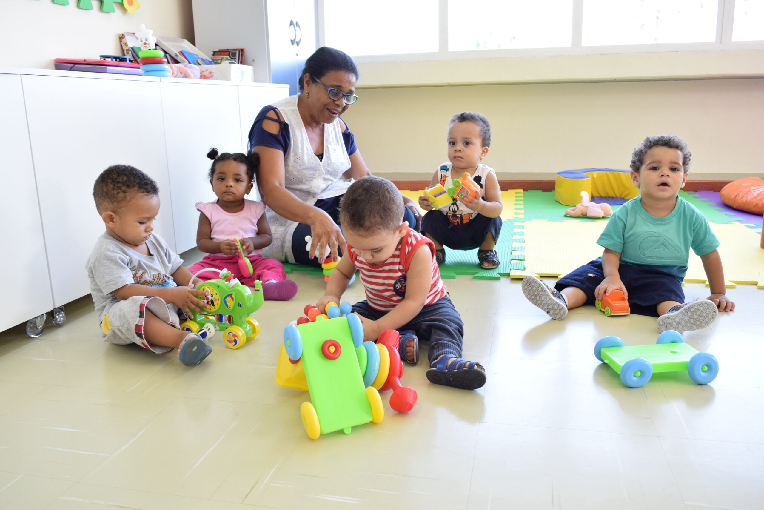 Professora de Educação Infantil sentada no chão da sala com cinco bebês sentados a sua volta brincando.