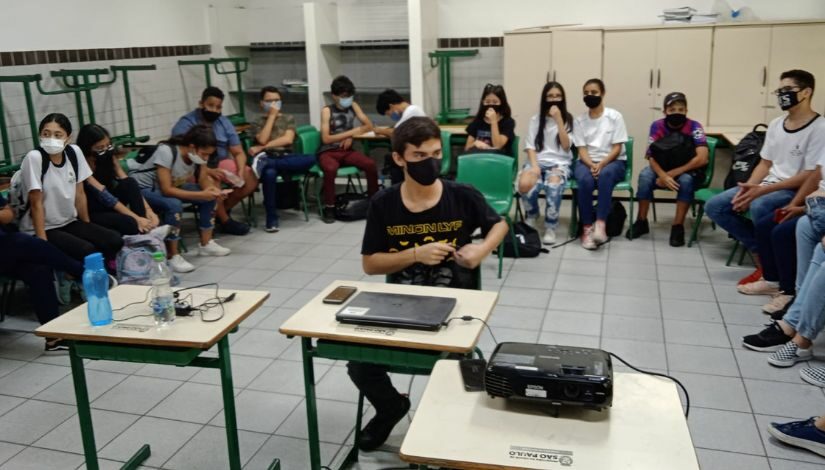 Sala de aula com estudantes sentados em suas cadeiras dispostas em círculo. No centro à frente, um rapaz de camiseta preta, sentado à sua mesa com um computador portátil e um celular sobre ela.