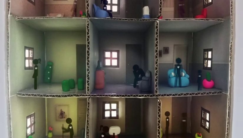 Imagem mostra uma caixa de papelão com várias divisórias formando quadrados menores. Dentro deles mini móveis e pessoas feitas de animação.