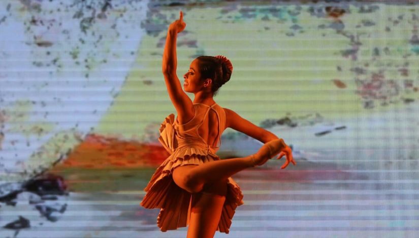 Fotografia de uma bailarina, ela está de costas, com o braço esquerdo levantado e o direito esticado ao lado. Uma de suas pernas está esticada atrás e ela usa um vestido na cor bege.