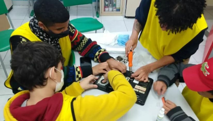 Fotografia de quatro estudantes em volta da mesa desmontando um computador portátil. Eles usam colete amarelo e estudam na Escola Municipal de Educação Bilíngue para Surdos (EMEBS) Anne Sullivan .