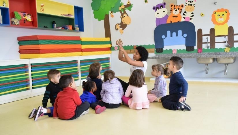 Professora de Educação Infantil sentada no chão. Ela está contando uma história para as crianças pequenas que estão sentadas ao seu redor, as crianças olham para sua mão gesticulando no ar acima da sua cabeça.