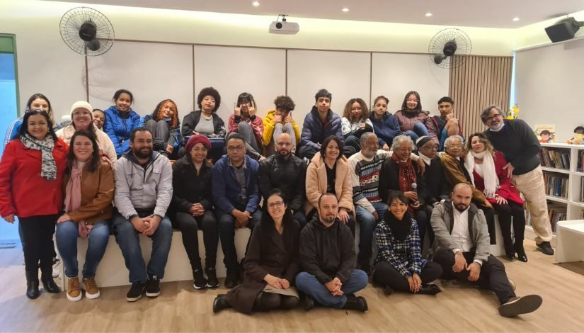 30 pessoas estão sentadas na Sala de Leitura, eles estão em uma arquibancada com duas fileiras, dentre elas, 4 pessoas estão sentadas a frente no chão. 