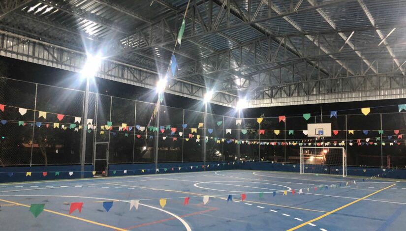Fotografia de uma quadra poliesportiva coberta, enfeitada com bandeirinhas coloridas. É noite e iluminação está ligada.