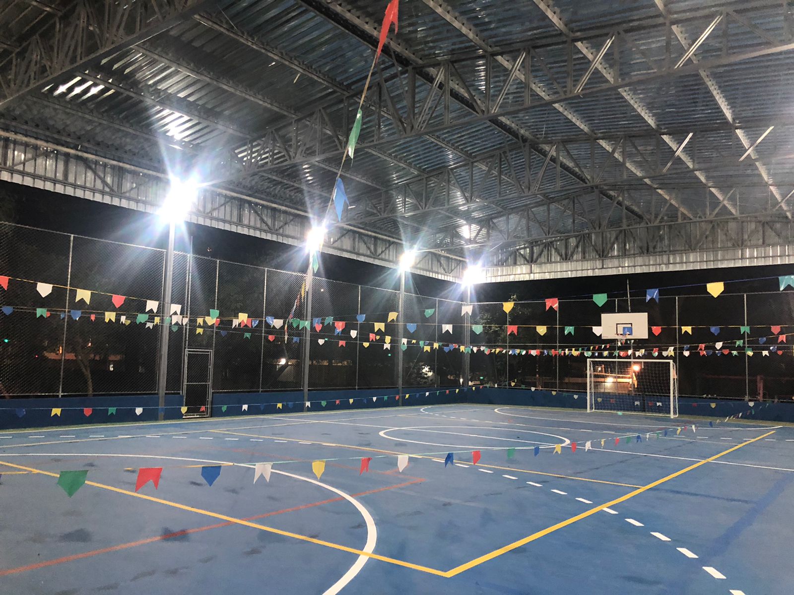 Fotografia de uma quadra poliesportiva coberta, enfeitada com bandeirinhas coloridas. É noite e iluminação está ligada.