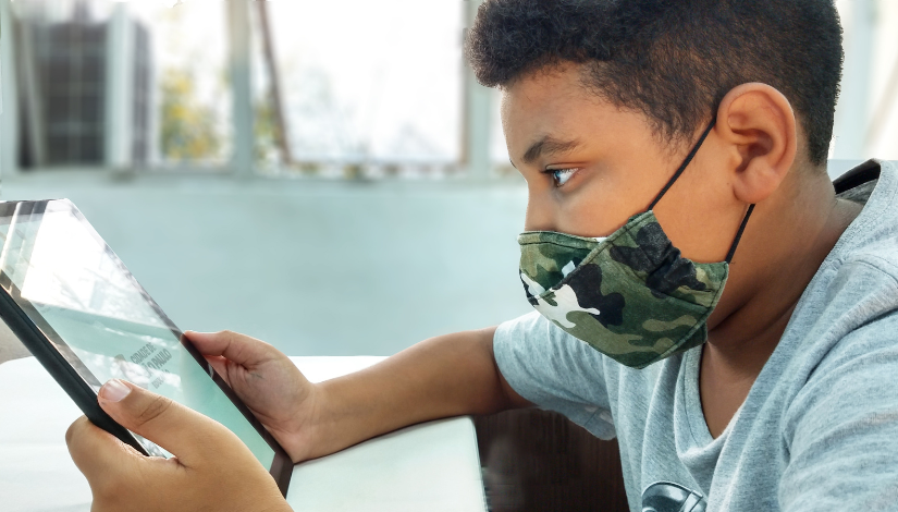 Fotografia tira de perfil de um menino que está segurando um tablet. Ele usa camiseta cinza e máscara. O tablet é preto e na tela há a logotipo da Secretaria Municipal de Educação.