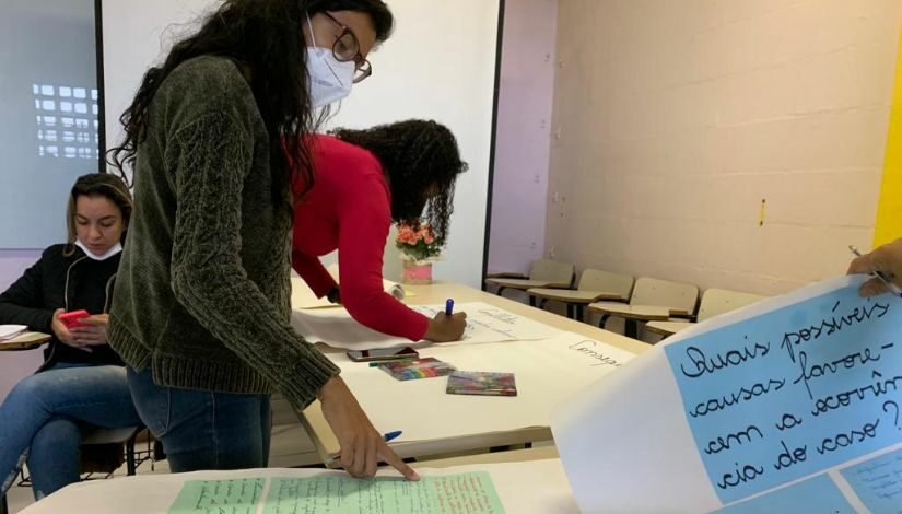 Fotografia de uma sala de aula com mesas e cadeiras. Duas mulheres em pé, uma escreve no cartaz sobre a mesa e a outra lê um cartaz diferente que também está sobre a mesa. No quadrante inferior direito, um cartaz escrito 'Quais possíveis causas favorecem a ocorrência do caso?'.