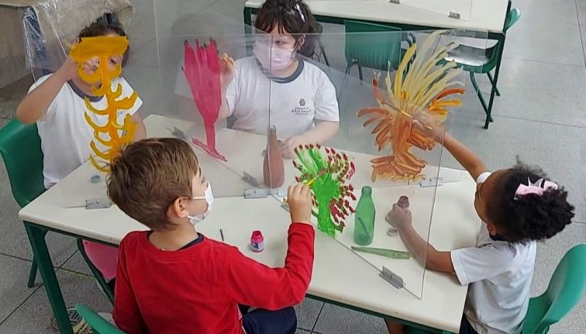 Quatro crianças pintando árvores coloridas em um suporte de acrílico posicionado sobre a mesa de forma vertical.