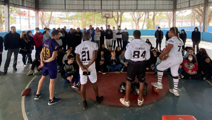 Fotografia de quatro jogadores de futebol americano conversando com um grupo de estudantes e educadores na quadra coberta da escola.