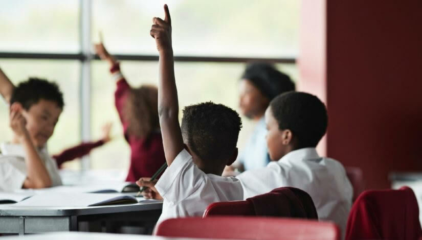Crianças em sala de aula, uma delas em destaque levantando a mão afim de fazer uma pergunta.