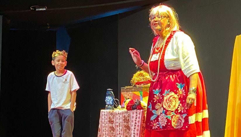 Estudante com a camiseta do uniforme e uma coroa dourada na cabeça ao lado de uma mesa cheia de objetos como baú, lamparina e tecidos, e uma contadora de histórias que veste blusa branca e jardineira vermelha com flores.