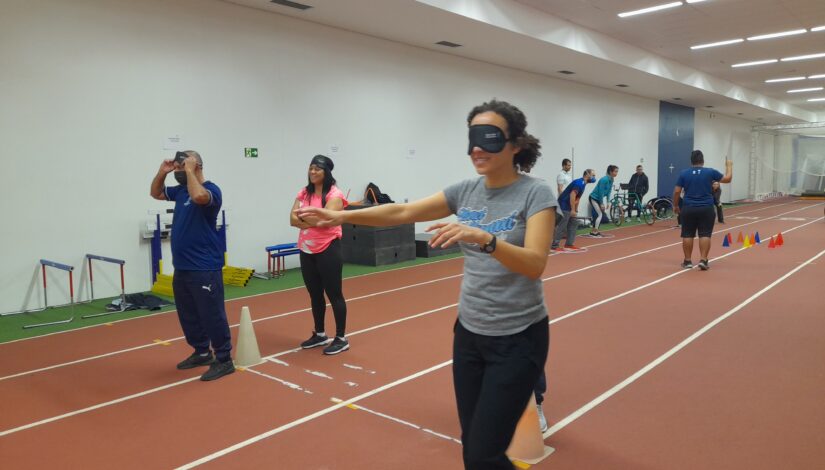 A imagem mostra professores caminhando vendados pela pista de atletismo do Centro Paralímpico Brasileiro. O chão é laranja, com listras brancas separando as pistas e cones coloridos, além de outros acessórios de atletismo. Na frente, uma mulher de cabelo cacheado curto, calça preta e camiseta cinza caminha vendada com os braços erguidos para frente. Ao lado direito dela estão um homem e uma mulher que também participam da atividade, com as vendas erguidas. Ao fundo estão outras seis pessoas envolvidas em outras atividades.