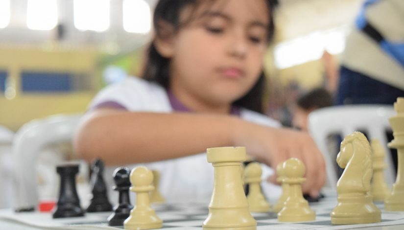 Menina olha para tabuleiro de xadrez e movimenta uma peça com sua mão direita.