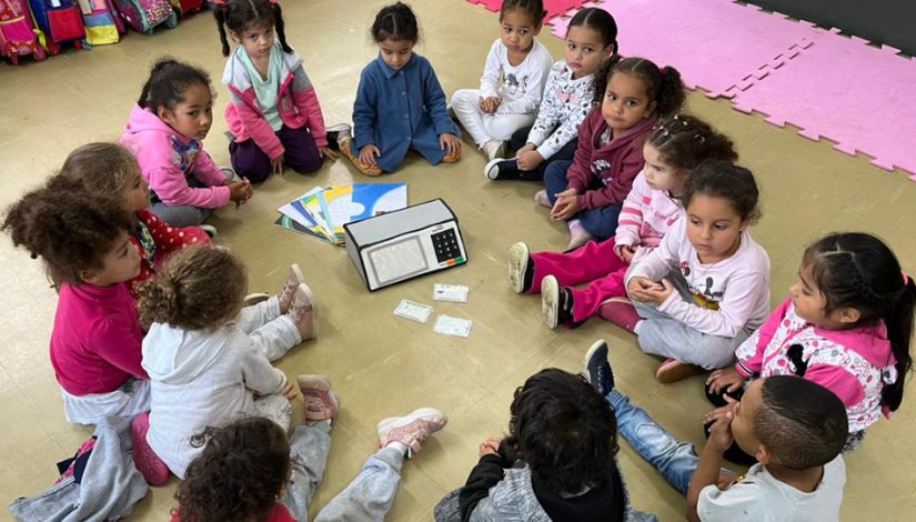 Crianças pequenas sentadas em roda com livros e urna eletrônica no meio.