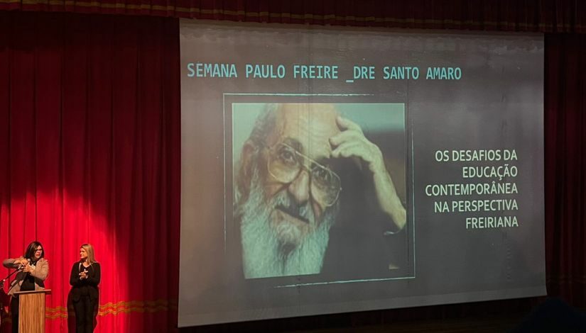 Fotografia de um palco com cortinas vermelhas e um telão com a imagem de Paulo Freire onde se lê 