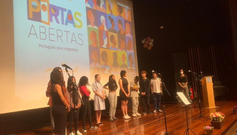 Fotografia de um palco com 11 pessoas uma ao lado da outra e ao fundo projetado no telão está escrito "Portas Abertas - Português para Imigrantes".