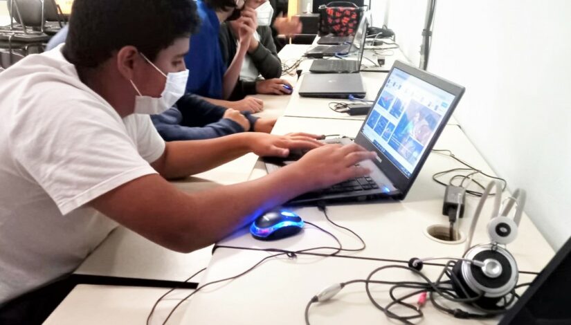 Fotografia mostra 5 estudantes que estão sentados à frente de notebooks. Na mesa vemos mouses, fios e headset.