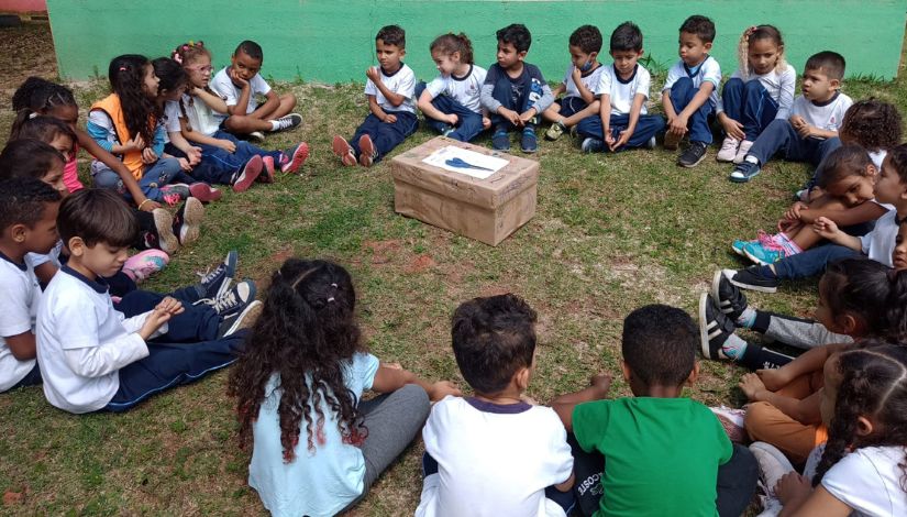26 crianças estão sentadas no gramado, em círculo. Elas estão olhando para caixa que está no centro da roda, que está forrada com um papel marrom e com desenhos a sua volta.