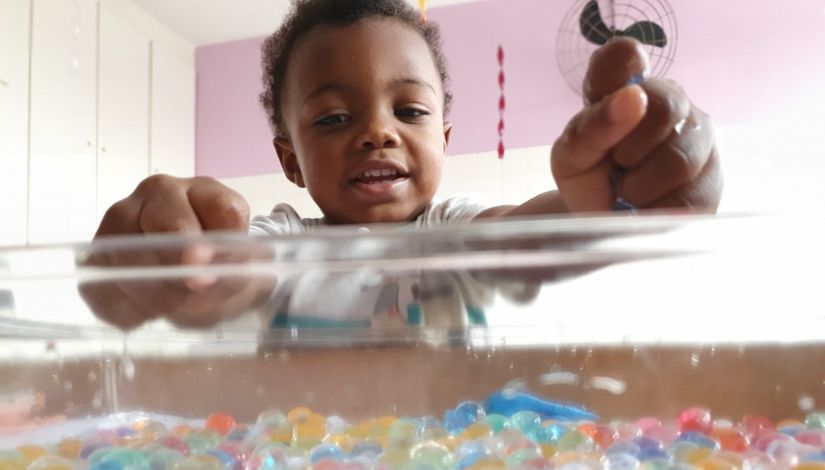 Criança está de pé brincando com recipiente transparente que está na sua frente. Dentro dele, há diversas bolinhas coloridas e água. O menino está com a mão fechada, segurando algumas das bolinhas.