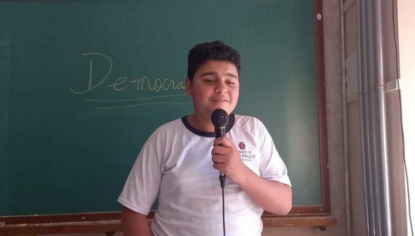 Um estudante está de pé e segura um microfone. ele está de costas para uma lousa que está escrito "democracia".