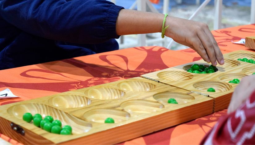 Fotografia de um tabuleiro de mancala de madeira com peças verdes e uma mão pegando as peças.