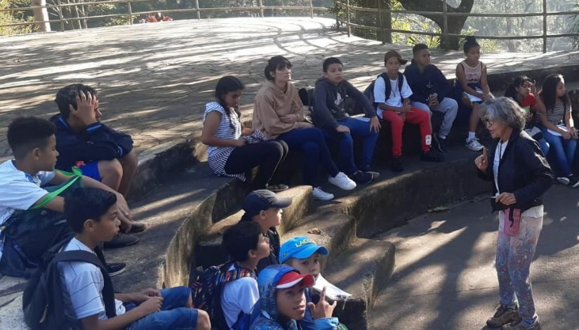 Estudantes estão sentados em degraus no Parque do Carmo, eles estão atentos, olhando para frente, onde há uma mulher de pé que está explicando para eles.