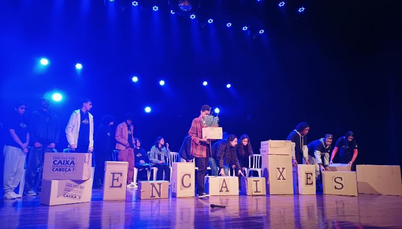 Estudantes estão em cima de um palco onde tem cadeiras e caixas. Cada caixa está com uma letra na frente, que juntas formam a palavra "Encaixes", há uma caixa em cada lateral que está com frases escritas. Um menino está de pé no centro segurando uma caixa com uma planta dentro.