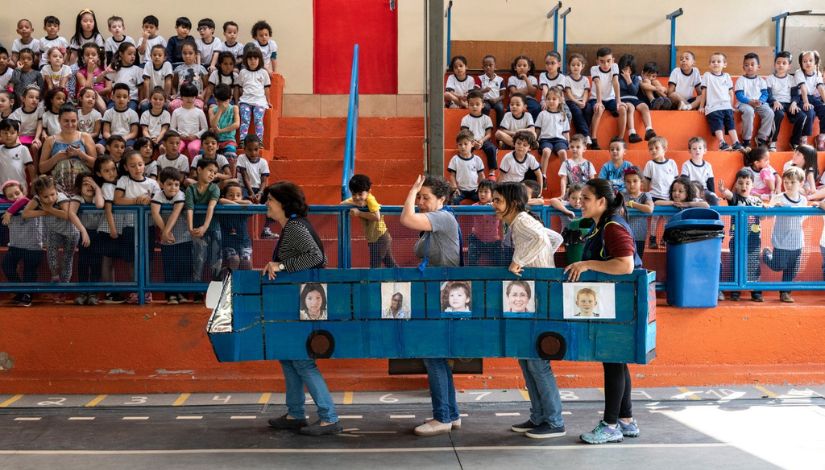 Fotografia de uma arquibancada com muitas crianças e pessoas adultas na quadra dentro de um ônibus feito de papelão.