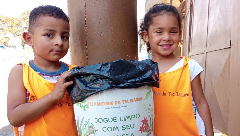 Um menino e uma menina estão com coletes laranjas escrito "CEI - Cantinho da Tia Isaura". Entre eles há uma lixeira que eles confeccionaram, ela está com um saco de lixo e com uma arte escrito "CEI Cantinho Da Tia Isaura, Cuidando do mundo da gente" "Jogue limpo com seu planeta".