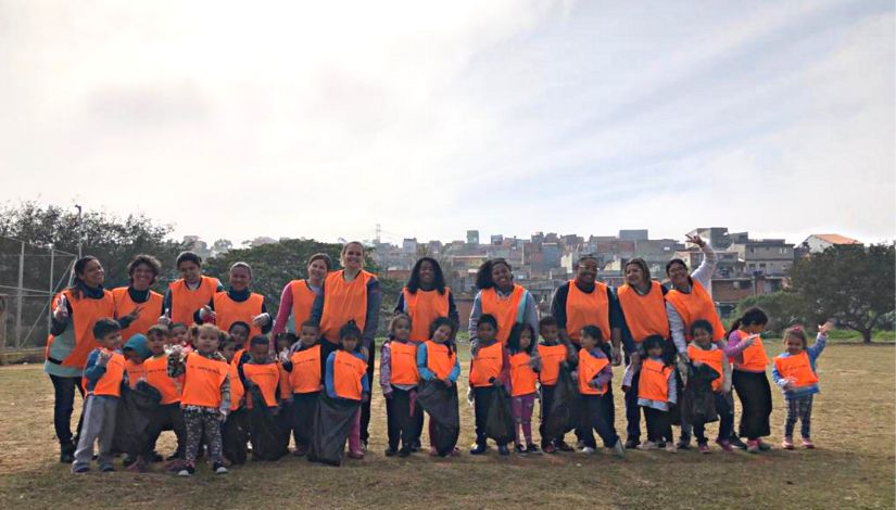 Fotografia mostra 11 adultos e 21 crianças posicionadas à frente deles. Eles estão usando coletes laranjas e segurando sacos de lixo pretos.