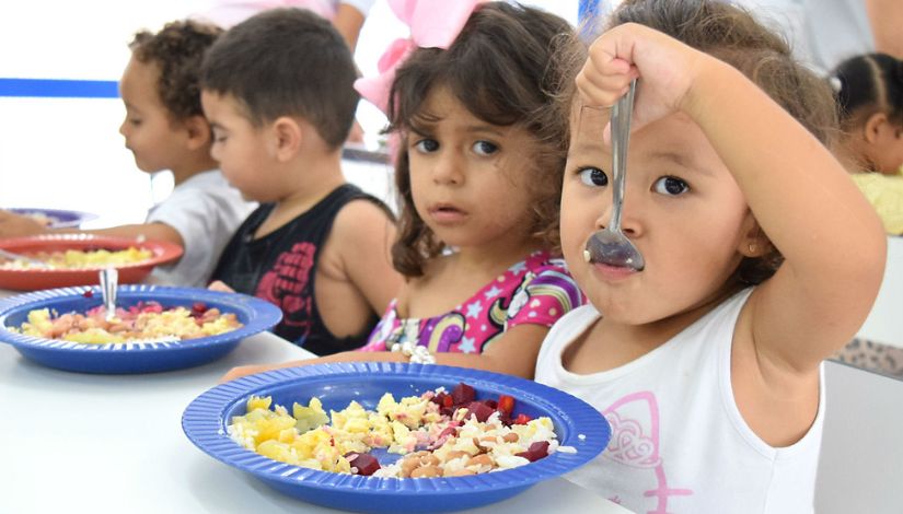 Crianças sentada à mesa fazendo sua refeição. Em primeiro plano, uma menina coloca a colher na boca, em sua frente está o prato com arroz, feijão, beterraba e outros legumes.