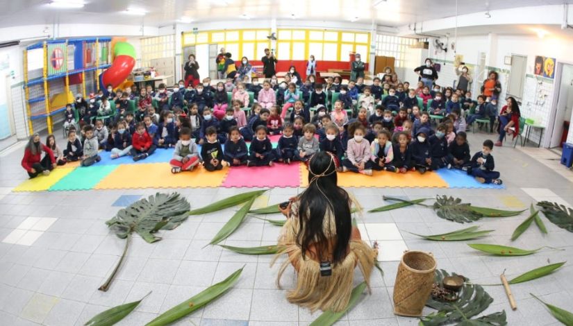 Um grupo de crianças, todas sentadas em tatames no chão, observam uma apresentação de uma mulher com vestes indígenas e folhas no chão.