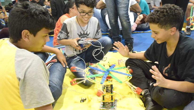 Fotografia com três meninos montando um robô com peças de robótica. Um deles segura um controle remoto e aciona um botão que faz uma roda girar, os outros dois olham atentamente para o movimento do robô.