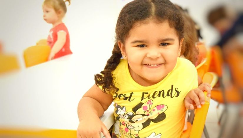 Fotografia de uma criança sorrindo. Ela olha para a câmera e usa camiseta amarela escrito 