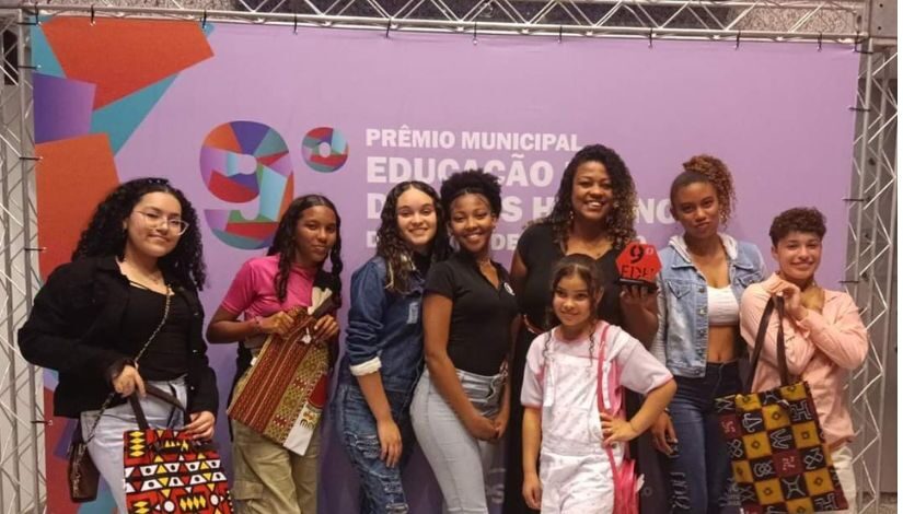 Na foto há 7 mulheres e 1 criança, elas estão de pé olhando para a foto e seguram sacolas com estampas étnicas. Uma delas é a professora, que segura o trófeu do 9° Prêmio de Educação em Direitos Humanos. Ele é hexagonal, vermelho e está escrito 9º EDH. 