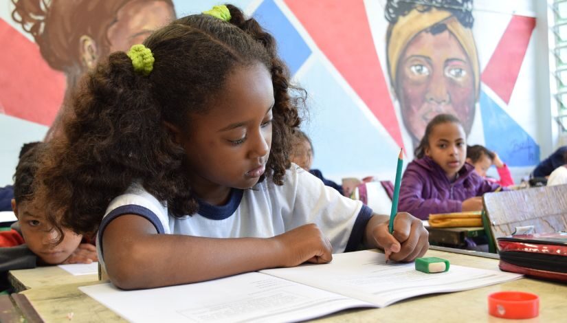 Fotografia de uma estudante negra, ela está concentrada escrevendo no livro sobre sua mesa.