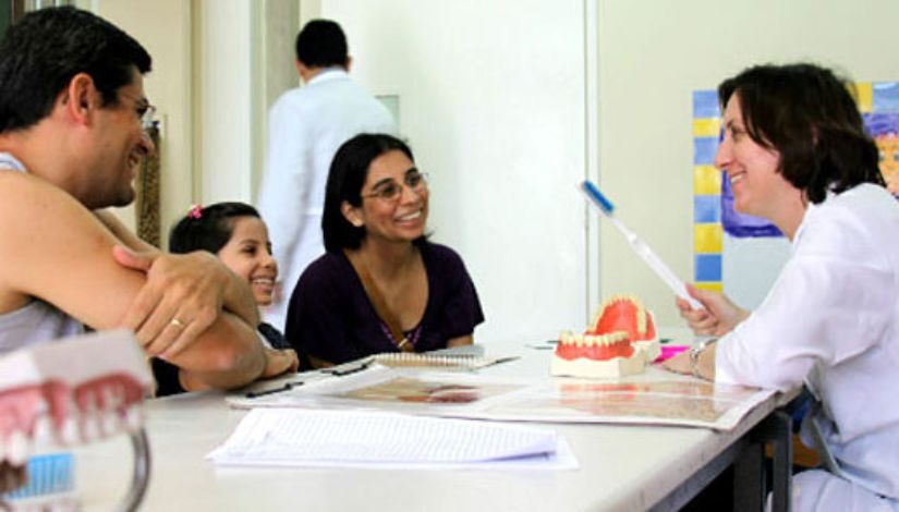 Fotofrafia de uma pessoa com jaleco branco segurando uma escova de dentes gigantes e conversando com uma criança e duas pessoas adultas ao lado dela. Todos estão sorrindo e sentados à mesa que também possui uma dentadura gigante sobre ela.