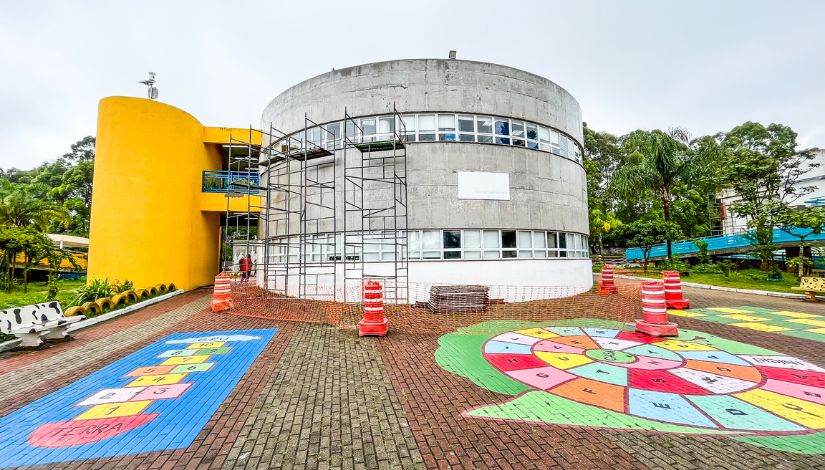 Fotografia de um prédio arredondado de um CEU, o chão está pintado com amarelinha e um desenho colorido de um caracol.